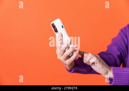 This image captures an elderly adult woman in a purple sweater interacting with a modern smartphone against a vivid orange backdrop. The focus is on h Stock Photo