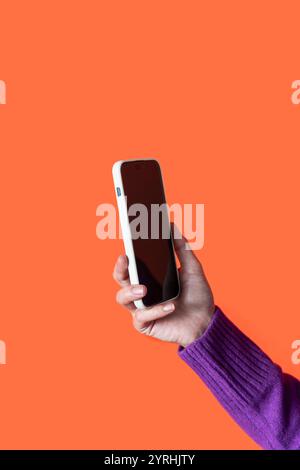 A female hand in a purple sweater holds a white smartphone, with a blank black screen, extended towards the camera against a vibrant orange backdrop Stock Photo