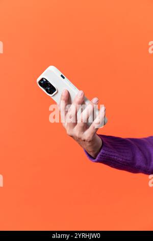 A close-up view of a Caucasian adult woman's hand holding a white smartphone with triple cameras, set against a vibrant orange backdrop. The focus is Stock Photo