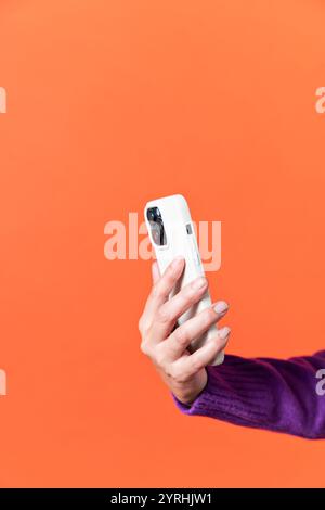 A close-up image of a female hand holding a white smartphone with a triple camera. The background is a vibrant, solid orange color, emphasizing modern Stock Photo