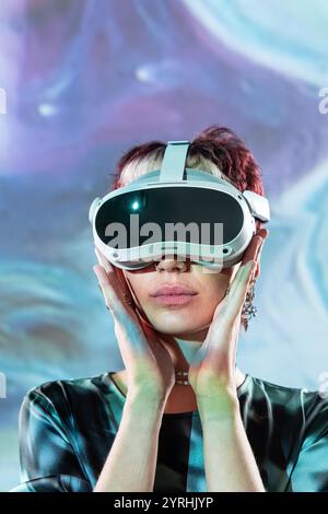 A young woman interacts with cutting-edge virtual reality technology, her hands gently framing a sophisticated VR headset against a vivid, abstract ba Stock Photo
