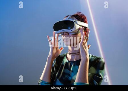 A young woman interacts with cutting-edge virtual reality technology, her hands gently framing a sophisticated VR headset against a vivid, abstract ba Stock Photo