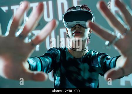 A young woman, sporting stylish attire and multiple accessories, engages with a digital VR universe, exploring simulated environments hand-forward and Stock Photo