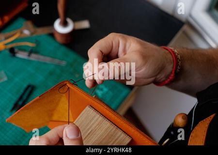 A craftsman meticulously stitches leather by hand, showcasing the art of leather working in a workshop Tools and materials are arranged on the craftin Stock Photo