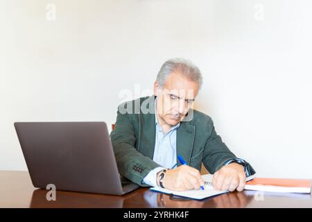 Elderly businessman focusing on paperwork at his desk, with a laptop nearby. Wearing a green blazer, he is deeply engaged in his work in a bright offi Stock Photo