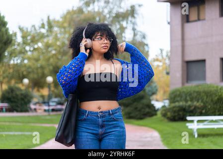 African American girl with afro hair, wearing headphones, walks through a university campus She is in casual attire, showcasing a vibrant and dynamic Stock Photo