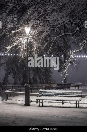 A tranquil winter evening scene in Geneva, Switzerland Snow blankets a park bench under a softly glowing streetlamp, evoking a peaceful, serene atmosp Stock Photo