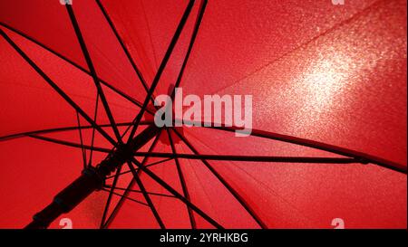 Detailed view of a red umbrella seen from below, showcasing its intricate frame and fabric texture as sunlight filters through, creating a vibrant glo Stock Photo