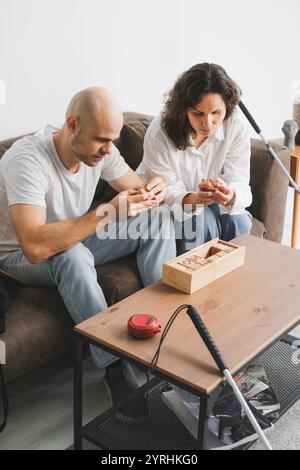 A blind couple sits on a couch, concentrating on solving a wooden puzzle on a coffee table. A white cane rests nearby, indicating attention to tactile Stock Photo