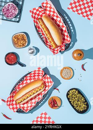 A flat-lay of two classic hot dogs in buns with mustard, arranged with various toppings and condiments on a vibrant blue background Stock Photo