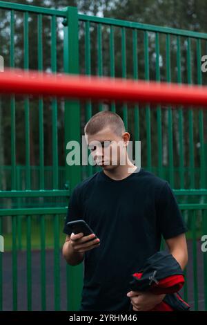 A young man stands in front of a green metal fence, looking intently at his smartphone. He holds a jacket and appears to be in a park or outdoor area, Stock Photo