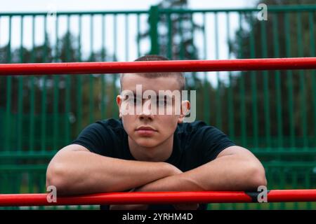 A young man in a black shirt leans thoughtfully on a red bar at an outdoor sports park. The green fence and trees create a serene background, capturin Stock Photo