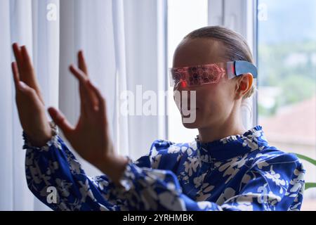 A woman in a blue floral blouse interacts with augmented reality using hi-tech, glowing glasses, standing by a window. The scene conveys innovation an Stock Photo