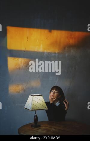 A woman sits at a wooden table with a lit lamp, casting warm shadows on the gray wall. The scene evokes a sense of contemplation and tranquility. Stock Photo