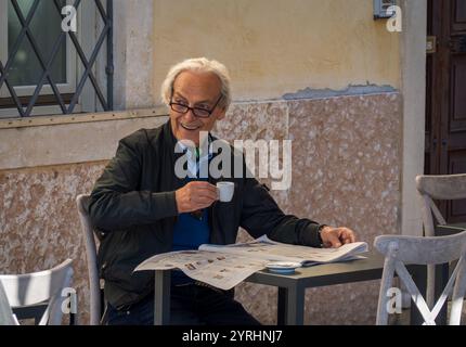 Verona, Italy - November 10, 2024: Smiling elderly man sipping espresso while reading newspaper at outdoor cafe table. Relaxed and content atmosphere with natural lighting and casual setting. Stock Photo