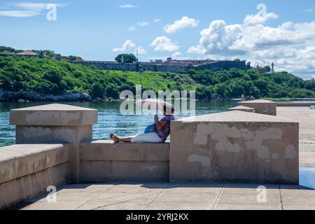 A Woman resting under the sun with an umbrella at Castllo de San Salvador de la Punta and Castillo de San Carlos de la Cabana in the horizon, La Haban Stock Photo