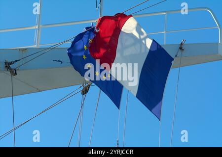 Close-up of the European Union and French flags on a ship, with the EU flag showing some wear and tear. The flags flutter against a bright, cloudless Stock Photo