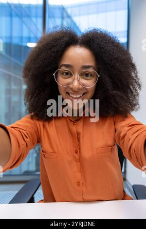 Young woman businesswoman in an orange blouse taking a creative selfie at the office. Her joyful expression and natural charisma shine through the camera lens. Perfect for themes of empowerment Stock Photo
