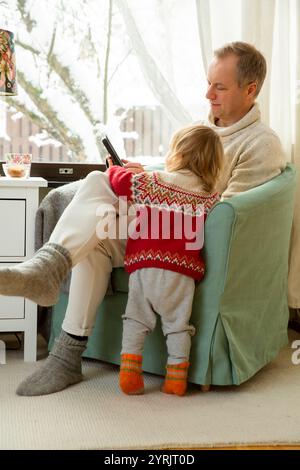 Middle age man and toddler sitting home in a chair by window with with scenic winter view of trees in snow. Family using tablet together.  Stock Photo
