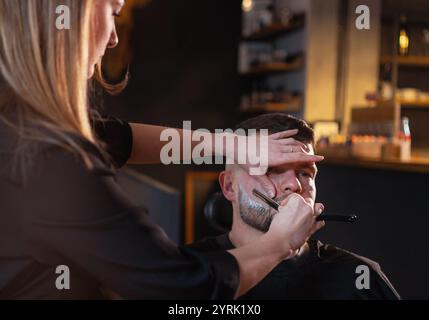 Long-haired young female Barber gently shaving bearded man face bristle while he in chair. Modern low light black style barber shop interior. Hair and Stock Photo
