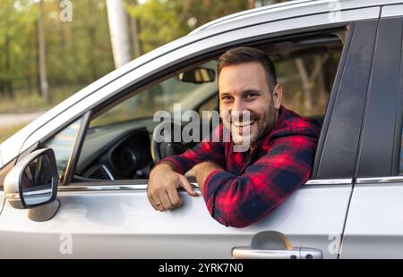 Portrait of handsome smiling man looking through window on driver seat in car standing in rural landscape Stock Photo