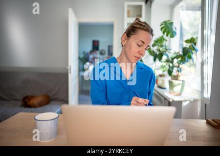 Thoughtful businesswoman works on laptop, taking notes in notebook in calm home office. Remote work Stock Photo