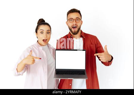 Excited man and woman, holding laptop with a blank screen, pointing at it with amazement and enthusiasm, promoting content or product, dressed casually, stand on isolated white background Stock Photo