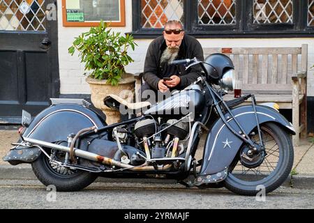 A parked 1940 black Indian Chief motorcycle, with the rider sitting on a bench having a drink and looking at his mobile. Stock Photo