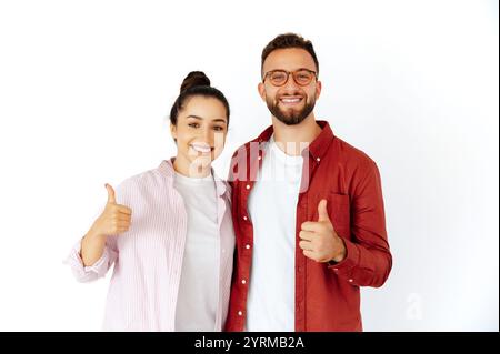 Smiling man and woman, arabian or caucasian ethnicity, dressed casually, standing together, showing thumbs up as a gesture of approval and positivity, standing on isolated white background Stock Photo