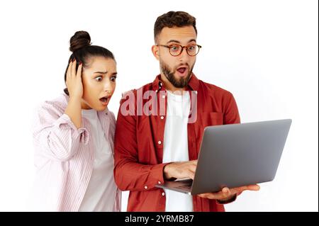 Shocked man and woman of arabian or caucasian ethnicity, looking at a laptop screen, expressing disbelief and amazement, dressed casually, standing on isolated white background, intense emotions Stock Photo