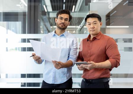 Two businessmen stand in a modern office holding documents and a tablet. They engage in a business discussion, focused on strategy and collaboration. T Stock Photo