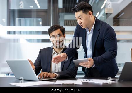 Two business professionals engage in a collaborative discussion in a modern office environment. One stands holding a tablet, while the other works on a laptop. The setting highlights teamwork. Stock Photo