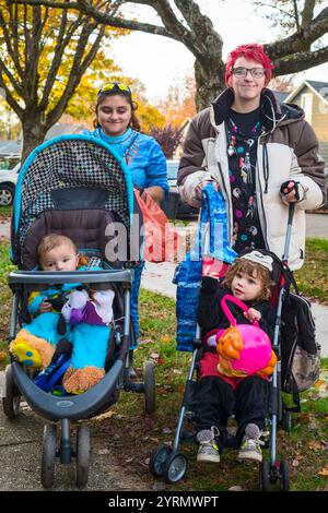 Two mothers push their children down the street in halloween costumes posing for photographer on a working class neighborhood street in the midwestern. Stock Photo