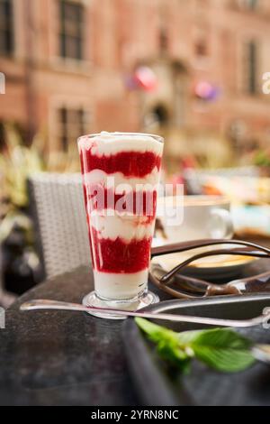 A delicious dessert layered with creamy white and vibrant red layers, served in a tall glass on a table with a mint garnish and a cup in the backgroun Stock Photo