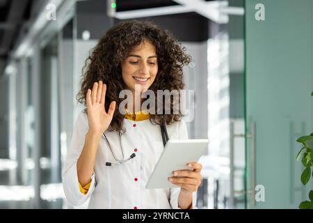 A smiling medical professional engages in a virtual consultation using tablet. The doctor, with a stethoscope around her neck, waves she interacts online, showcasing approach to healthcare. Stock Photo