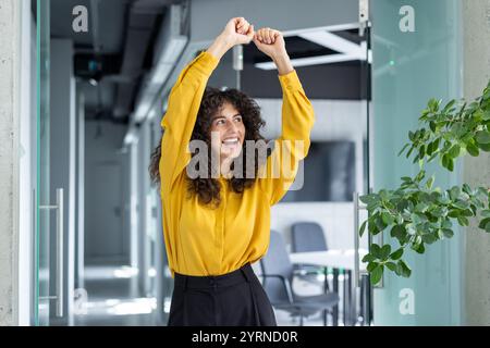 A joyful young woman celebrates with raised arms in a bright office. Her expression conveys excitement and accomplishment, epitomizing workplace success and positive energy. Stock Photo