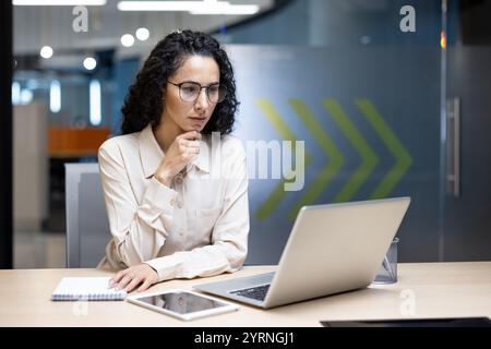 A businesswoman deeply focused on her laptop, surrounded by office essentials like a notebook and tablet. This setting captures a professional atmosphere, ideal for depicting determination Stock Photo