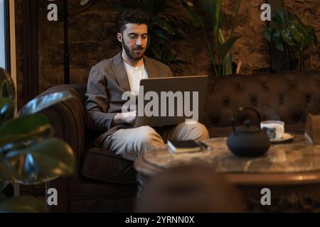 Professional man in casual blazer working concentrated on laptop in modern cafe. Stone wall texture and indoor plants create distinctive coworking atm Stock Photo