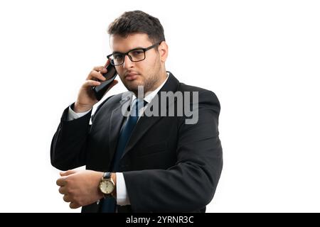 Serious businessman in smart casual suit and glasses talking on phone call showing wrist watch as timekeeping corporate concept isolated on white back Stock Photo