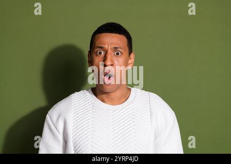 Surprised young man with wide eyes and open mouth wearing a white sweater stands against a khaki background Stock Photo