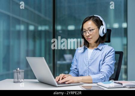 Asian woman wearing headphones works diligently on a laptop at a desk. Her focused expression shows deep concentration on her task. The office suggests productivity and modern technology usage. Stock Photo