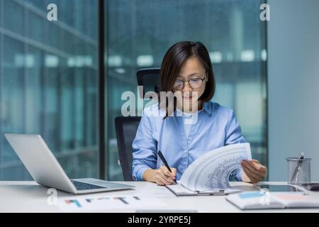 Asian woman concentrates on analyzing data at her office desk. She reviews documents while using a laptop, exemplifying professionalism and focus in a modern workspace. Stock Photo