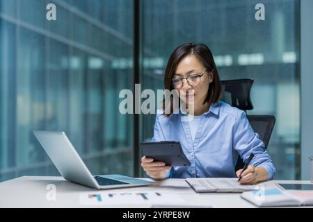 An Asian woman is deeply engaged in accounting work at a modern office desk. She is using a tablet and laptop to manage financial tasks efficiently. Her serious expression reflects concentration Stock Photo
