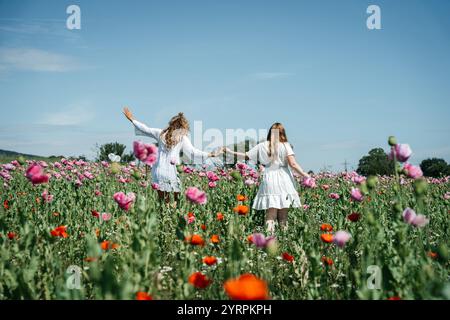 Two girls in white dresses embrace from behind, standing in a field of pink poppies gently swaying in the wind. A high-quality photo capturing the Stock Photo