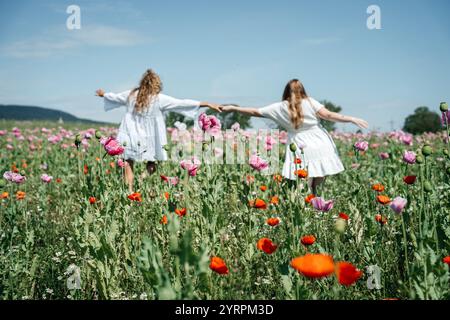 Two girls in white dresses embrace from behind, standing in a field of pink poppies gently swaying in the wind. A high-quality photo capturing the Stock Photo