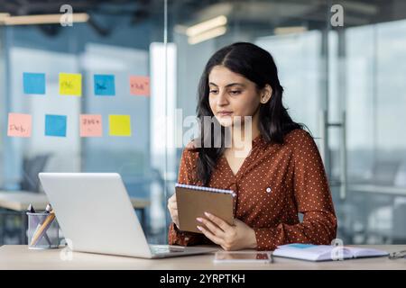A professional woman focuses on her laptop while taking notes in an office setting. She is engaged in online learning and working. Sticky notes on the glass hint at planning and organization. Stock Photo