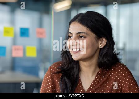 A young woman smiles warmly in a modern office setting. Colorful sticky notes on the glass wall add a creative touch, suggesting brainstorming and teamwork. Stock Photo