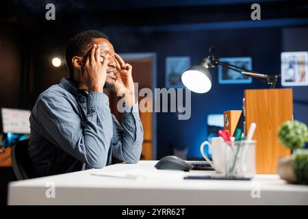 Male freelancer feeling overworked and yawning at home, trying to work under pressure to meet deadline. African american person studying online reports to help with business finances. Stock Photo
