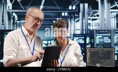 Older coworkers in data center, solving tasks on tablet while running scripts on laptop terminal. Experienced colleagues amidst servers, accessing databases and manipulating lines of code Stock Photo