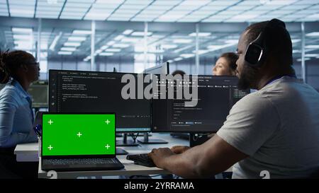 Isolated screen laptop next to programmer coding in server farm while listening music in headphones. Worker safeguarding hardware against unauthorized access with chroma key notebook, camera B Stock Photo